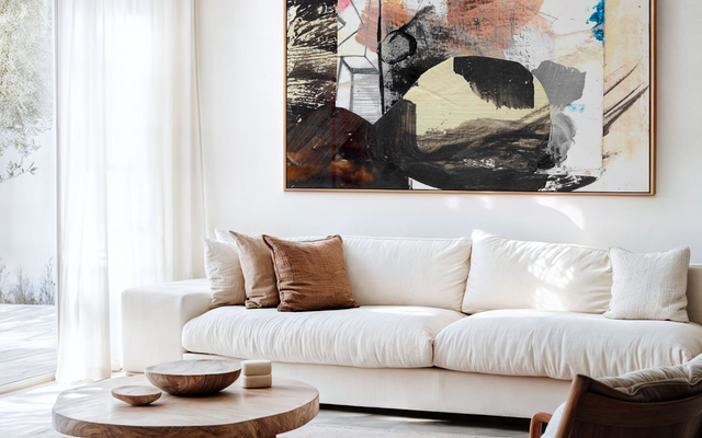 Sala de tons neutros com sofá branco com parede sobre o sofá exibindo um quadro decorativo em canvas, sem vidro, de arte abstrata em tons neutros.