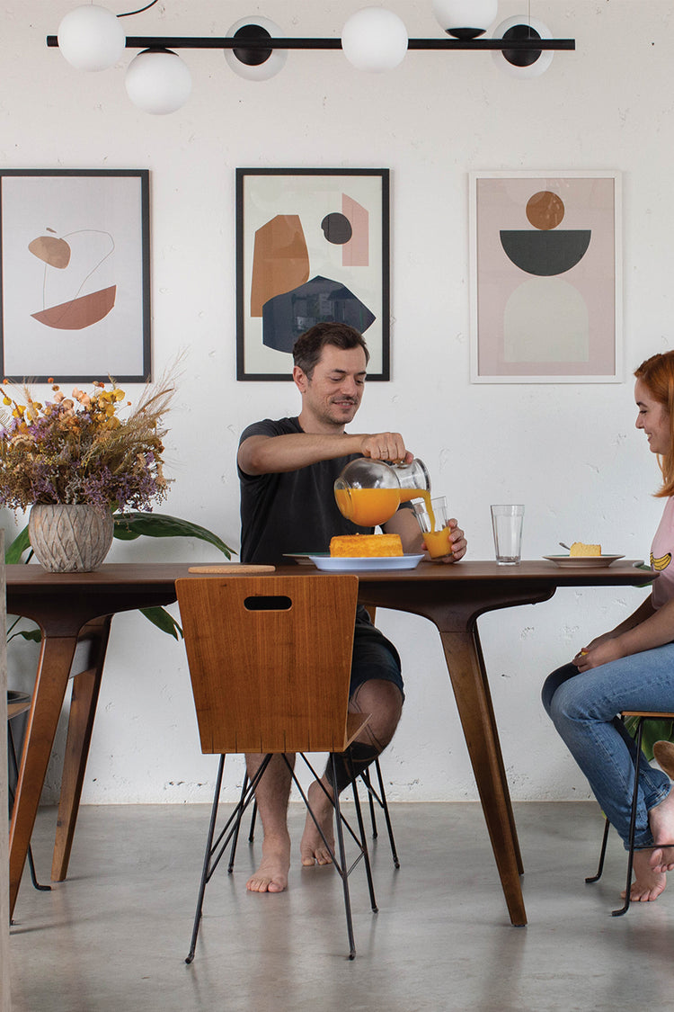 Casal tomando café da manhã em sala de jantar com decoração contemporânea, destacando três quadros com arte abstrata na parede. Mesa de madeira, flores secas e presença de um cachorro criam um clima leve e afetivo.