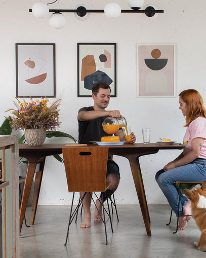 Casal tomando café da manhã em sala de jantar com decoração contemporânea, destacando três quadros com arte abstrata na parede. Mesa de madeira, flores secas e presença de um cachorro criam um clima leve e afetivo.