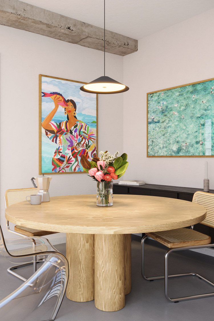 Sala de jantar com mesa redonda de madeira e cadeiras com estrutura metálica, decorada com pôster colorido de mulher tropical e arte fotográfica aérea de piscina, estilo contemporâneo com clima praiano e vibrante.