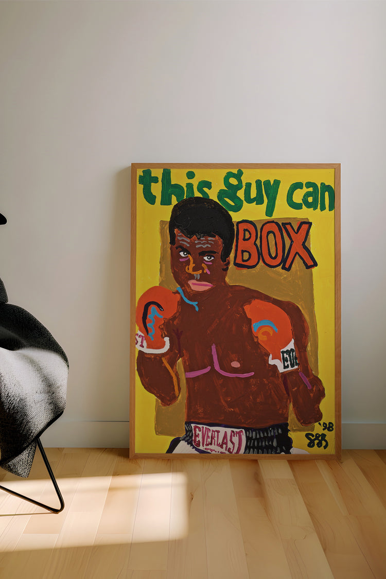 Quadro encostado na parede com ilustração colorida de boxeador em estilo pop e texto “this guy can box”. Obra vibrante com fundo amarelo e toques de humor em ambiente contemporâneo com piso de madeira.