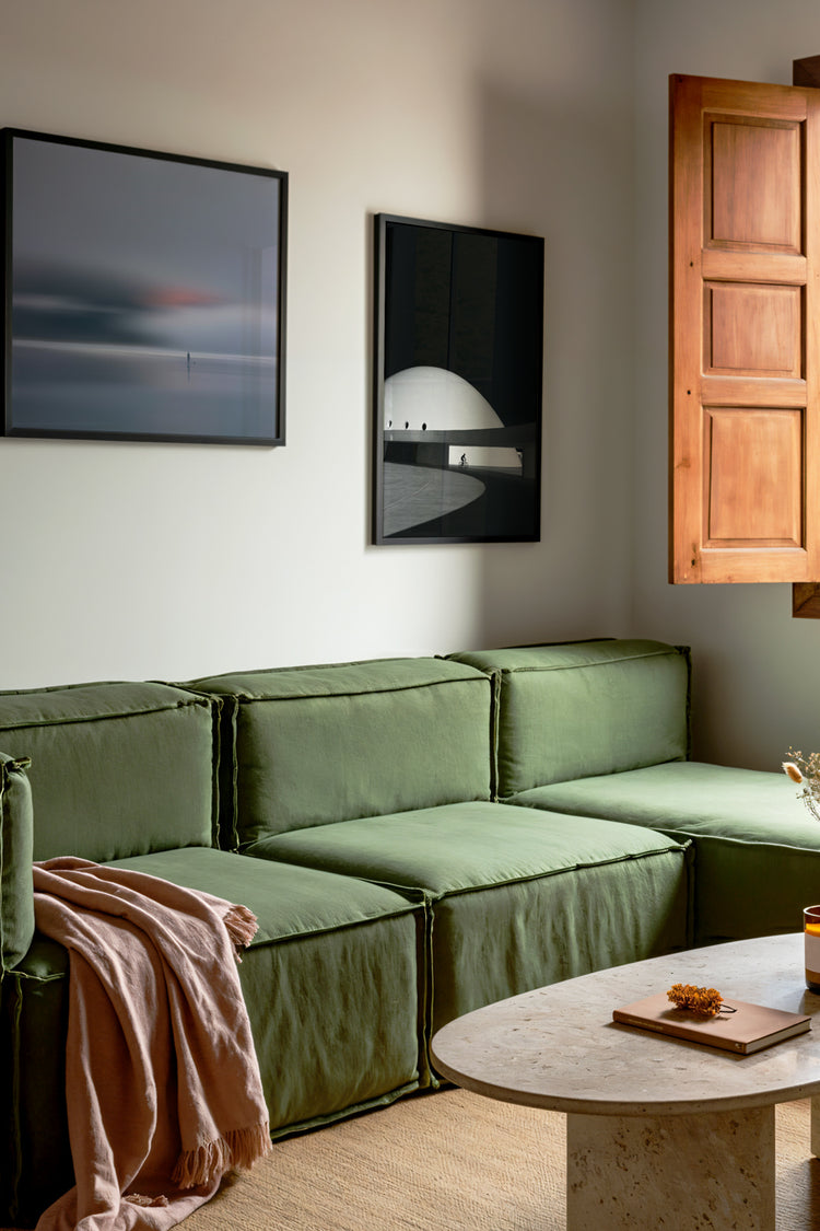 Sala de estar moderna com sofá verde de linho, manta bege casual, mesa de centro em mármore travertino, e decoração minimalista. Paredes com quadros decorativos — arte abstrata em tons suaves e fotografia em preto e branco com tema arquitetônico. Janela de madeira com luz natural destacando o design aconchegante e sofisticado. Decoração de interiores, design moderno, arte na decoração.