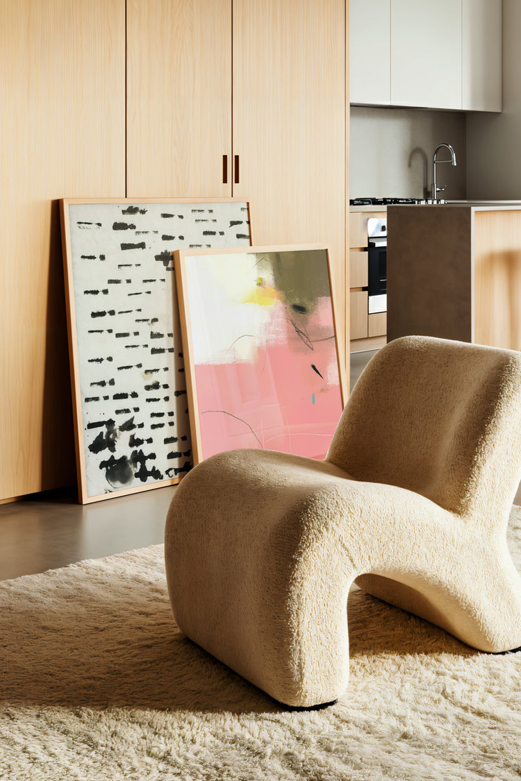Sala moderna com poltrona curvilínea em tecido bouclé bege, design icônico de mobiliário contemporâneo, destacando conforto e sofisticação. Tapete felpudo em tom neutro, reforçando o aconchego do ambiente. Dois quadros decorativos encostados na parede: arte abstrata em preto e branco e pintura em tons vibrantes de rosa e amarelo, valorizando o design de interiores com estilo e personalidade. Armários em madeira clara e cozinha integrada minimalista, criando um conceito aberto elegante. Decoração moderna, ar