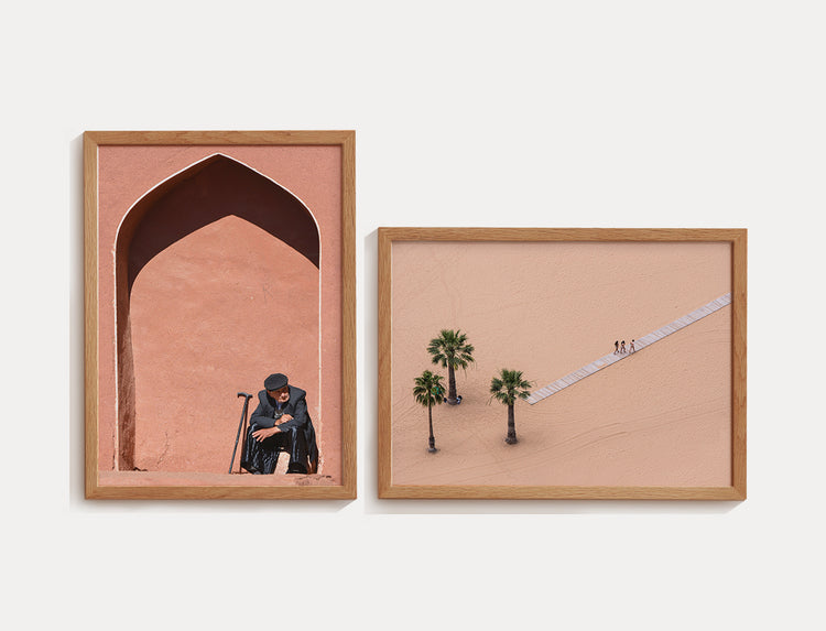 Duas fotografias emolduradas em madeira. A primeira mostra um homem idoso sentado sob um arco arquitetônico rosa, com bengalas ao lado. A segunda exibe uma vista aérea minimalista de um deserto claro com três palmeiras e uma passarela diagonal onde três pessoas caminham.