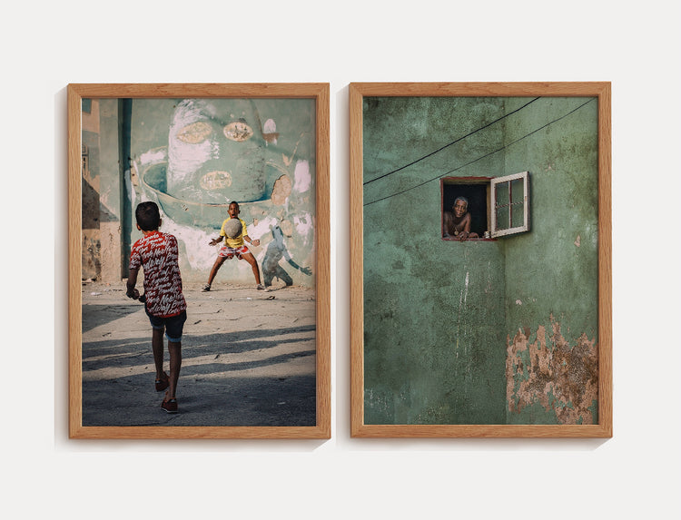 "Duas fotografias de rua com moldura de madeira. A primeira mostra duas crianças brincando em uma rua com uma parede grafitada ao fundo. A segunda retrata uma pessoa sorridente espiando por uma pequena janela em uma parede verde desgastada, com fios atravessando a cena