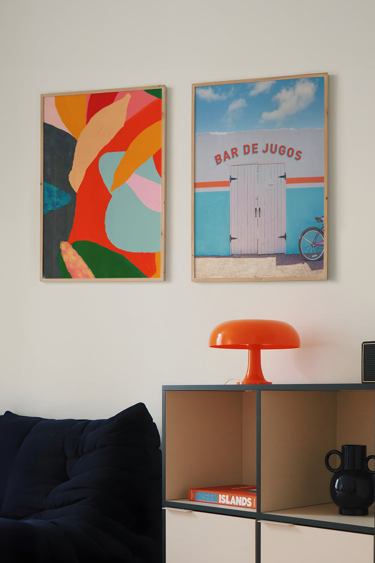 Parede decorada com pôster artístico colorido e fotografia de fachada tropical com bicicleta – decoração moderna com arte vibrante acima de sofá azul e luminária laranja retrô.