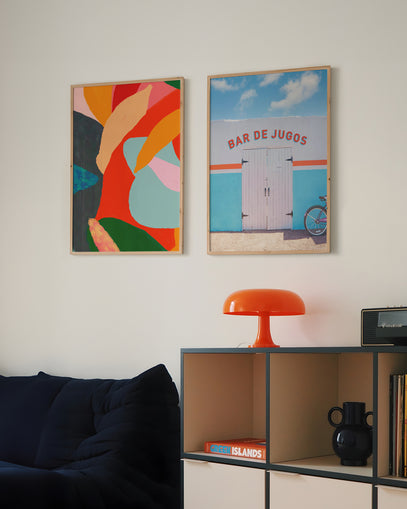 Parede decorada com pôster artístico colorido e fotografia de fachada tropical com bicicleta – decoração moderna com arte vibrante acima de sofá azul e luminária laranja retrô.