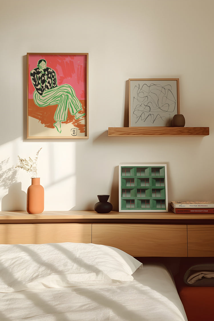 Quarto com decoração moderna e tons neutros, destacando três quadros sobre a cabeceira da cama. O principal, pendurado na parede, exibe uma figura feminina estilizada com roupas verdes e fundo vibrante em vermelho e rosa. Abaixo e ao lado, o outroo apresenta um formato quadrado com uma arte abstrata em riscos e o terceiro apresenta uma fotografia de um edifício em verde e complementam a estética artística do ambiente.