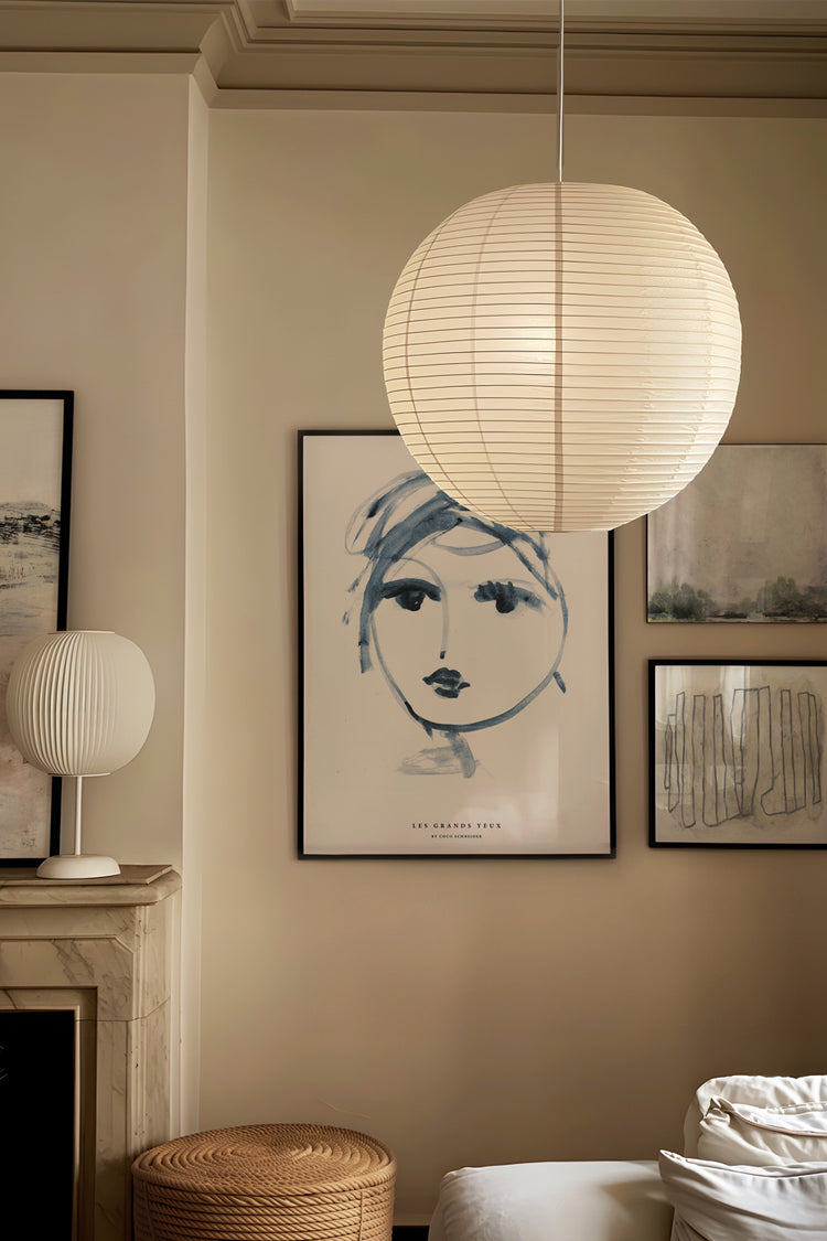 Ambiente sofisticado com iluminação suave, apresentando uma parede com quatro quadros artísticos. O destaque é um pôster central com ilustração de rosto feminino em traços azuis. Ao lado, obras em estilo abstrato com tons neutros compõem a galeria. Uma luminária pendente de papel e uma escultura luminosa sobre a lareira completam a decoração minimalista.