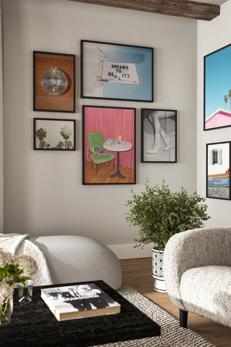 Sala descontraída com composição de quadros coloridos e estilos diversos, incluindo frases, fotografia, objetos e paisagens. Ambiente moderno com poltrona clara, mesa preta e plantas, transmitindo personalidade e criatividade.