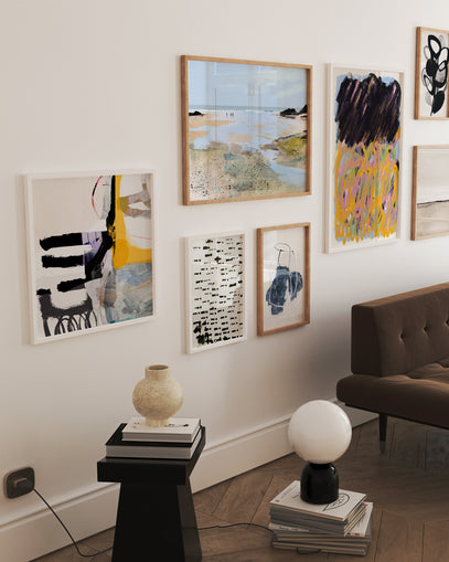 Composição de quadros variados em parede de sala com decoração contemporânea. As obras mesclam paisagens, arte abstrata e ilustração gráfica, criando uma galeria harmoniosa acima de sofá marrom.