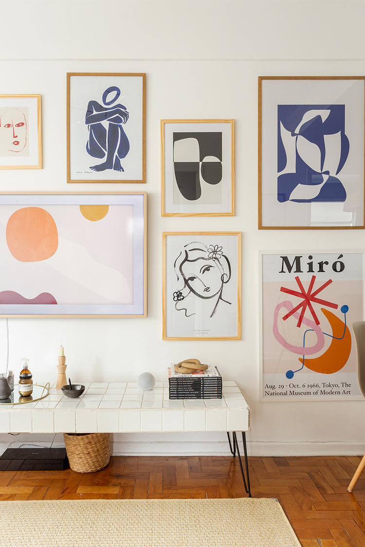 Parede com galeria de quadros decorativos modernos abstratos e ilustrativos, incluindo referências como Matisse e Miró. Ambiente claro e organizado com móveis minimalistas e piso de madeira.