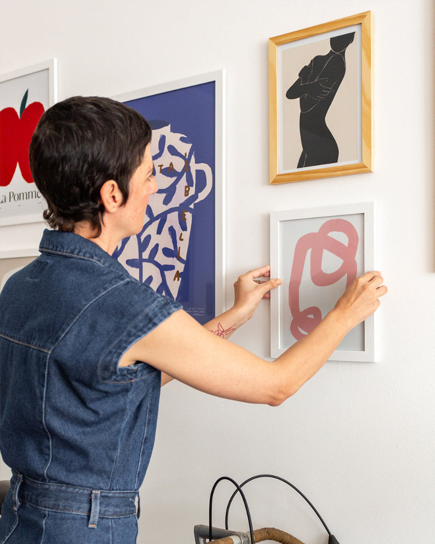 Pessoa ajustando quadro com arte abstrata em parede com composição de pôsteres modernos. Destaque para diferentes estilos de moldura e formas orgânicas em ambiente artístico e despojado.