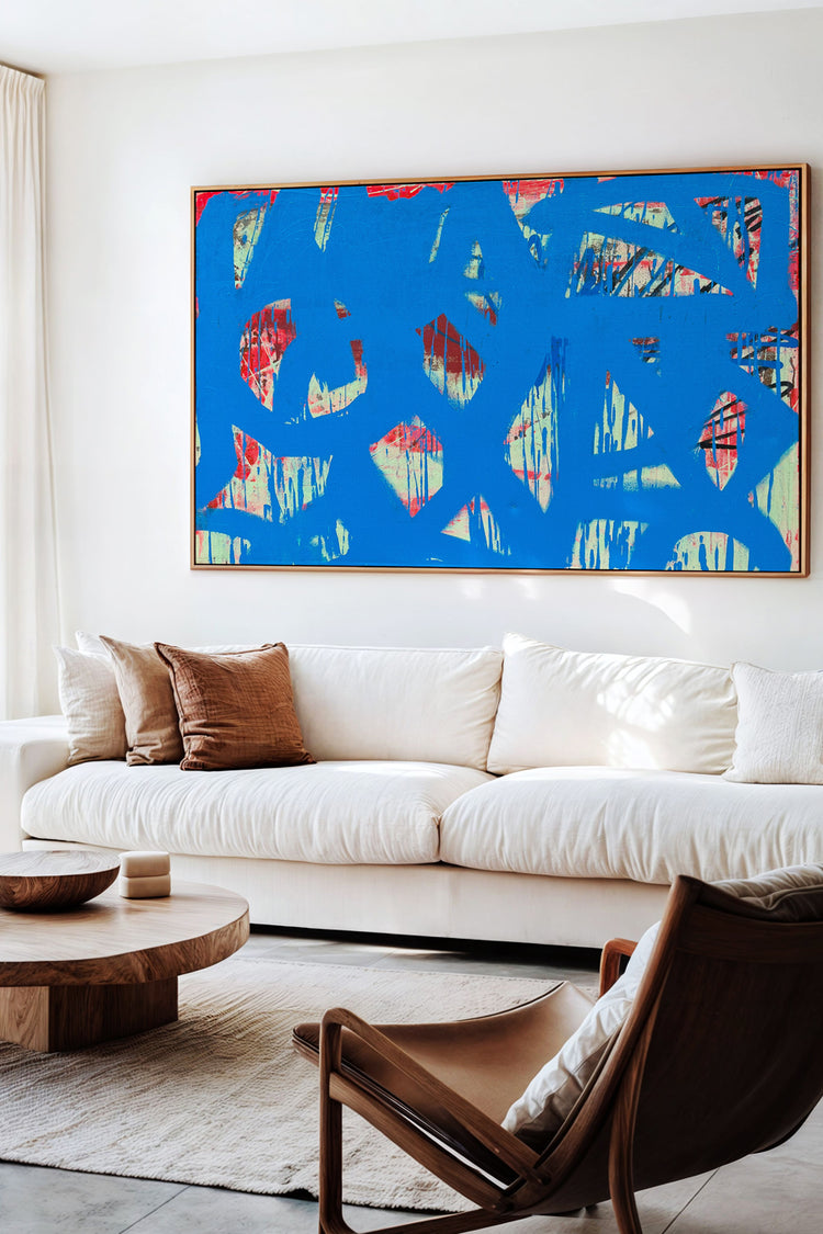 Sala de estar com decoração minimalista e tons neutros, destacando quadro grande com arte abstrata em azul vibrante. Composição elegante com sofá claro, mesa de centro de madeira e poltrona moderna.