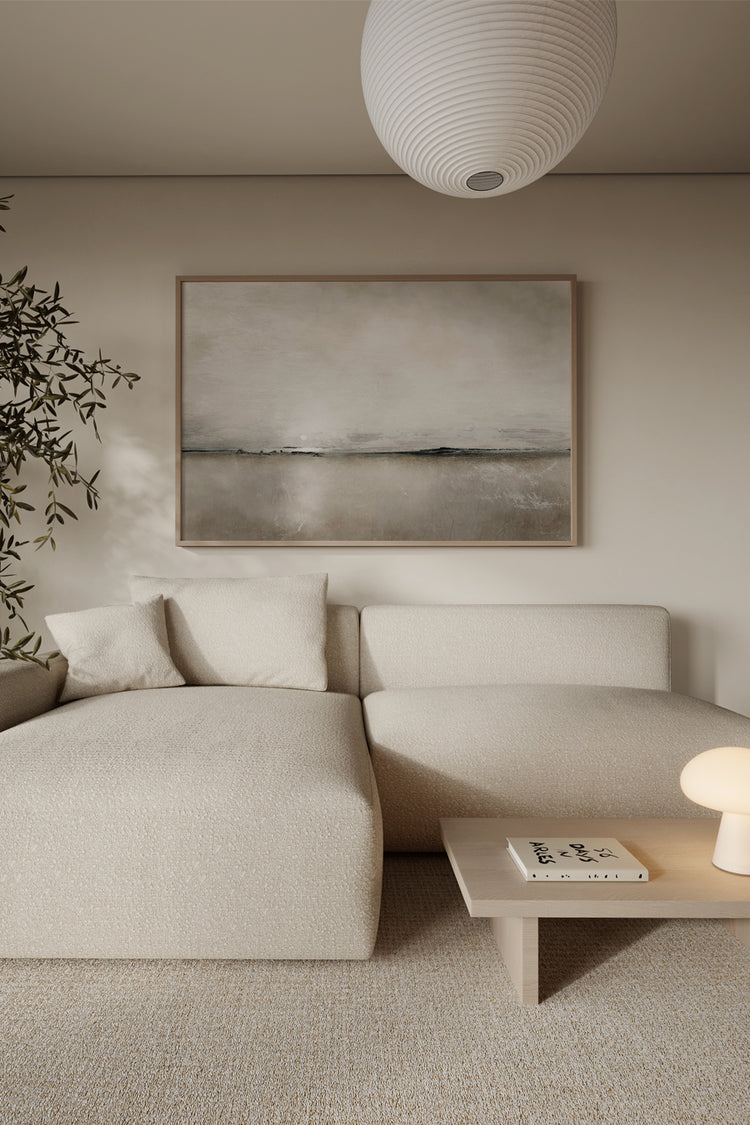 Sala de estar minimalista com quadro único, grande em tamanho 84,1x118,9cm, na moldura padrão mel. A Arte é minimalista e apresenta uma pintura abstrata de uma paisagem em tons neutos. Ao canto da sala há uma folhagem e no centro um abajur de formato de cogumelo na cor branca.