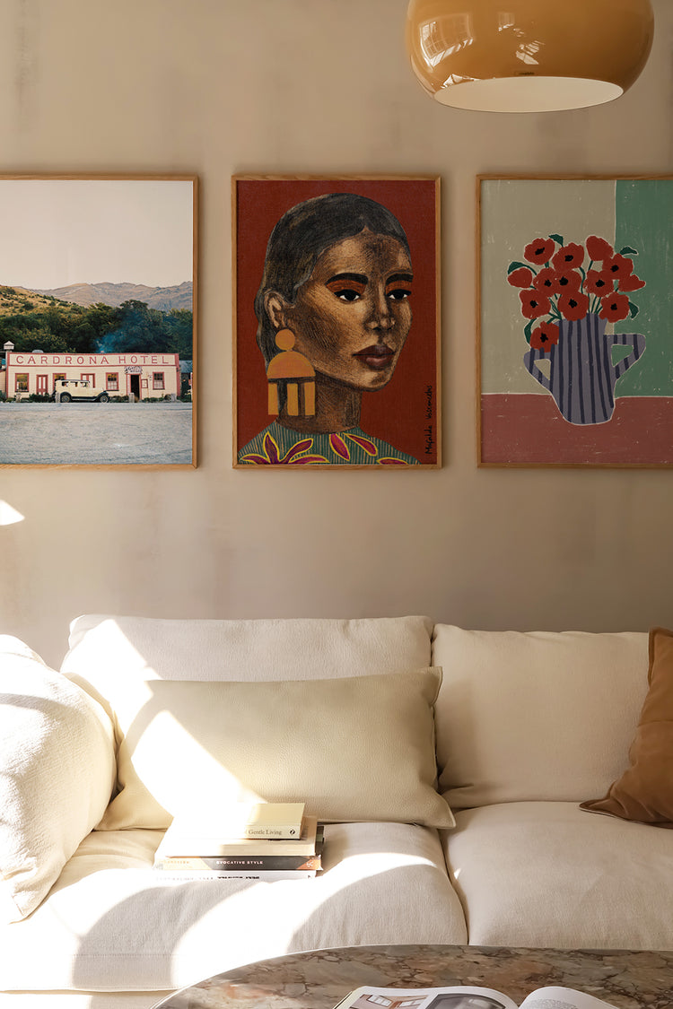 Trio de quadros com diferentes estilos artísticos em parede neutra sobre sofá off-white — fotografia de paisagem urbana, retrato feminino afro-descendente e natureza morta com flores, composição acolhedora e cheia de identidade cultural.