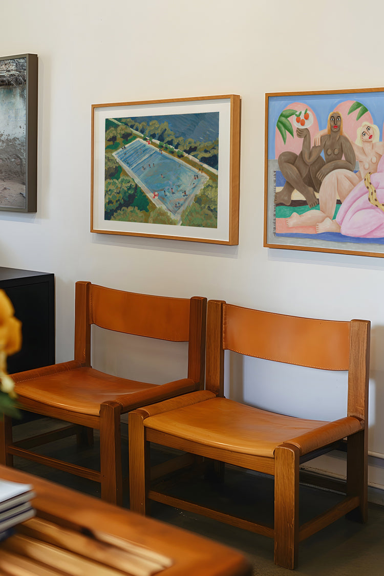 Parede com quadros contemporâneos coloridos em ambiente de galeria moderna — obras com temática tropical e feminina sobre cadeiras de couro natural e madeira clara, decoração artística e despojada.