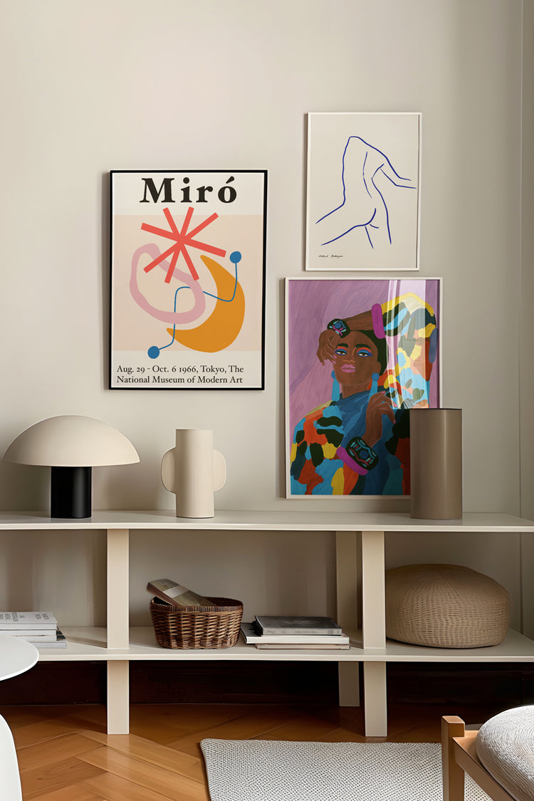 Ambiente moderno com parede decorada por pôster de Joan Miró, desenho minimalista de linha e ilustração colorida de figura feminina. Abaixo, aparador branco com objetos de design, livros e cestos, sobre piso de madeira em espinha de peixe.