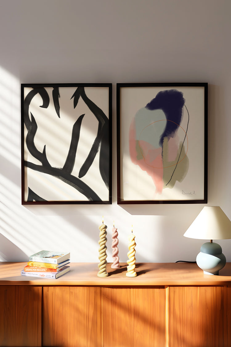 A imagem apresenta um ambiente acolhedor, com um aparador em madeira, com alguns objetos por cima, como abajur, livros e velas. Na parede acima, há dois quadros de moldura preta: um com a ilustração de um corpo feminino em linhas pretas pesadas e com fundo branco; e o segundo são manchas abstratas em tons de azul, rosa e verde sobre fundo branco. O ambiente possui uma luz solar que o torna mais aquecido e familiar.