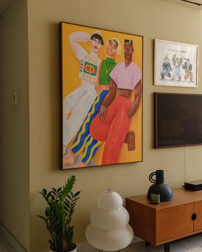 Parede amarela em sala de estar com móveis em cor de madeira. Na parede, há um quadro do artista Cépé, da Obrah, de três pessoas encostadas em uma parede. Ilustração despojada e colorida.