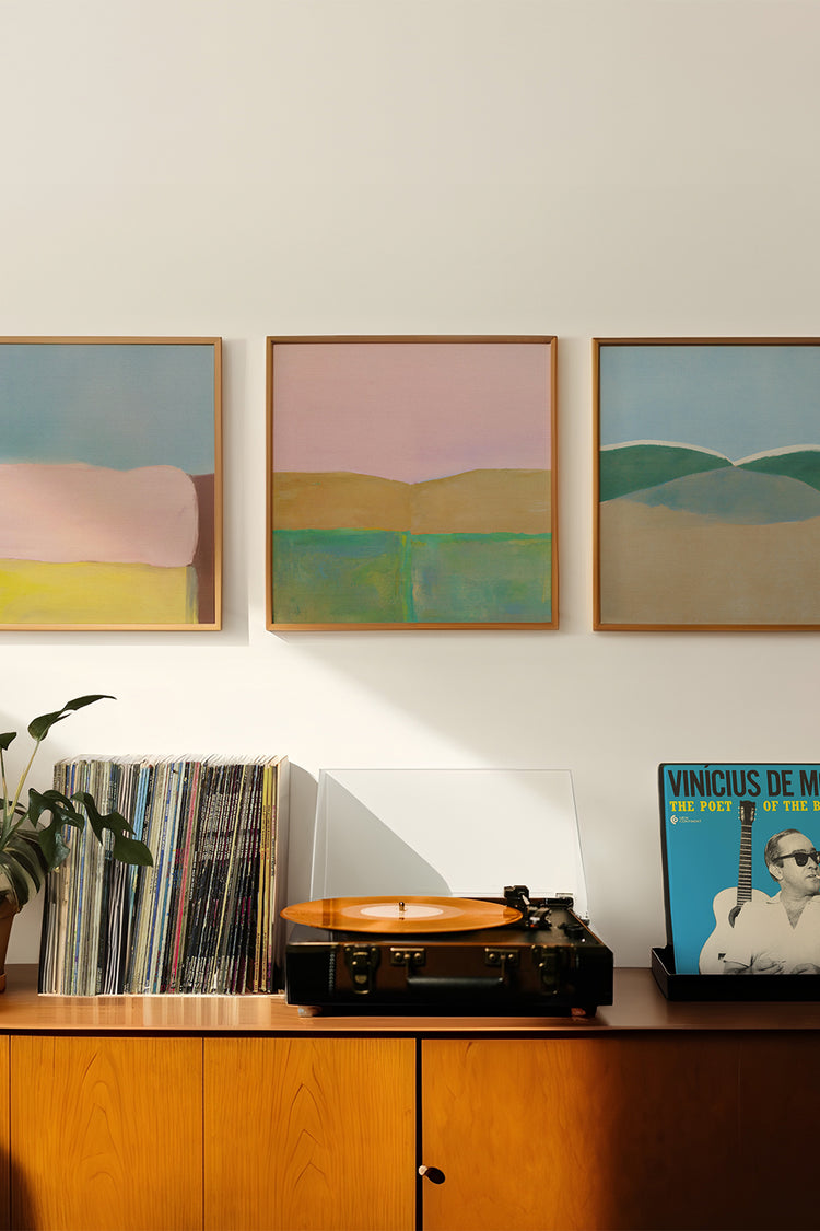 Trio de quadros abstratos em tons pastel sobre paisagens imaginárias, com composições em azul, rosa, verde e amarelo. Decorando parede acima de móvel de madeira clara com vitrola e discos de vinil, incluindo álbum de Vinicius de Moraes. Estilo moderno, aconchegante e sofisticado para salas de estar contemporâneas.