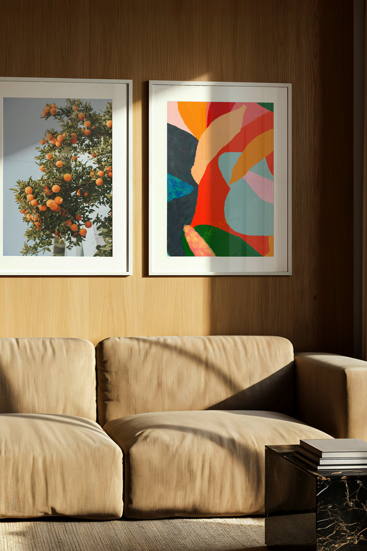 Sala de estar com decoração sofisticada, destacando sofá de veludo bege de design moderno, acompanhado por mesa lateral em mármore preto com detalhes dourados. Parede em painel de madeira com duas obras de arte emolduradas: fotografia botânica de árvore com frutas cítricas e arte abstrata colorida em tons vibrantes. Iluminação natural realçando texturas e cores, criando um ambiente acolhedor e elegante. Decoração de interiores, design contemporâneo, galeria de arte na parede, ambiente aconchegante e sofist