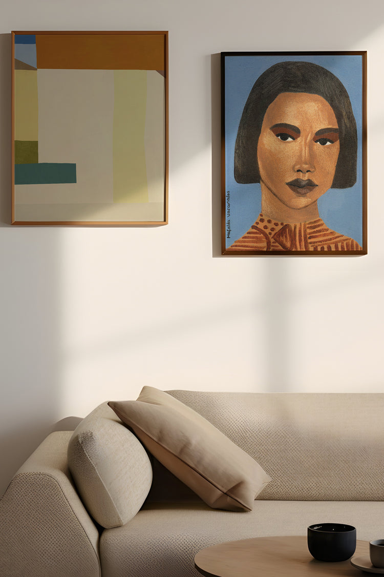 Dois quadros contemporâneos pendurados acima de sofá neutro, incluindo retrato feminino em fundo azul e pintura geométrica moderna. Composição artística para decoração sofisticada e atual.