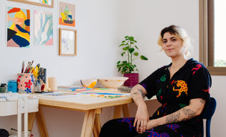 Artista Juline Lobão sentada em uma cadeira em seu studio com artes abstratacoloridas nas paredes e na mesa