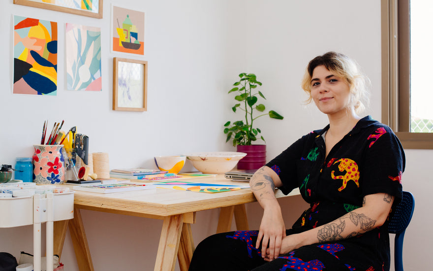 Artista Juline Lobão sentada em uma cadeira em seu studio com artes abstratacoloridas nas paredes e na mesa