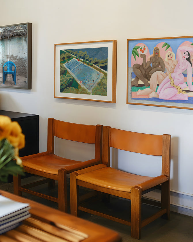 Parede com quadros contemporâneos coloridos em ambiente de galeria moderna — obras com temática tropical e feminina sobre cadeiras de couro natural e madeira clara, decoração artística e despojada.