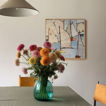 "Quadro com pintura abstrata náutica em tons claros na parede de sala de jantar com mesa posta, vaso verde de vidro com buquê de flores coloridas e cadeiras de madeira. Decoração artística e aconchegante com toque floral e marítimo.